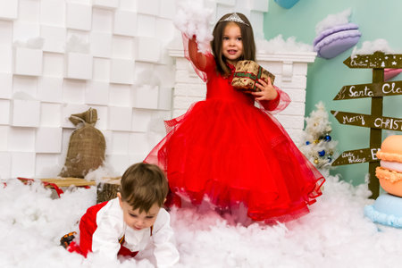 A little girl and a boy in red clothes have fun in the artificial snow by the fireplace in the studio. The girl is holding a box with gifts. Little kids in red christmas costume having fun beside a decorated christmas tree. Two kids playing with falling artificial snow flakes near a christmas tree.Kids playing with artificial snow flakes falling near a christmas tree. Christmas concept. High quality photoの写真素材