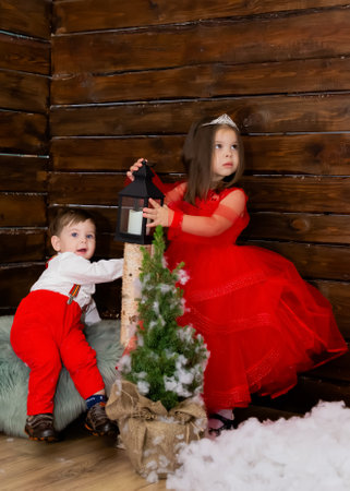 A little girl and a boy look at the Christmas candle. Wooden background. Preparing for the New Year and Christmas. A cheerful little girl looks at the Christmas lantern with a smile. Brother and sister in the red clothes at Christmas. Christmas concept. High quality photoの写真素材