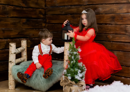 A little girl and a boy look at the Christmas candle. Wooden background. Preparing for the New Year and Christmas. A cheerful little girl looks at the Christmas lantern with a smile. Brother and sister in the red clothes at Christmas. Christmas concept. High quality photoの写真素材