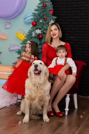 Family photo portrait. Mom and her two children and a white dog in red clothes celebrate the Chistmas, new year. Happy young mother and her daughter, son and a dog in a cozy dark living room on Christmas eve. Celebrating Xmas at home. Winter evening with family, kids and a dog. A little girl in a red dress is holding a gift. Family looking at camera holding many gifts boxes celebrating happy New Year. Merry Christmas presents. A child in a red dress holds a gift box. Christmas conceptの写真素材