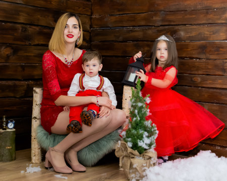 A little girl and a boy and their mother look at the Christmas candle. Wooden background. Preparing for the New Year and Christmas. A cheerful little girl, boy and woman looks at the Christmas lantern with a smile. Brother and sister and their mom in the red clothes at Christmas. Christmas concept. High quality photoの写真素材