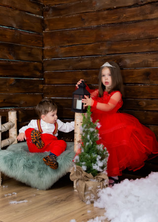 A little girl and a boy look at the Christmas candle. Wooden background. Preparing for the New Year and Christmas. A cheerful little girl looks at the Christmas lantern with a smile. Brother and sister in the red clothes at Christmas. Christmas concept. High quality photoの写真素材