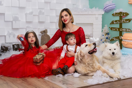 Family photo portrait. Mom and her two children and two white dogs in red clothes celebrate the Chistmas, new year. Happy young mother and her daughter, son and dogs in a cozy dark living room on Christmas eve. Celebrating Xmas at home. Winter evening with family, kids and dogs. A little girl in a red dress is holding a gift. Family looking at camera holding many gifts boxes celebrating happy New Year. Merry Christmas presents. A child in a red dress holds a gift box.Christmas conceptの写真素材