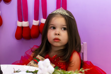 A little girl in a red dress is holding a gift. A little girl holds a gift on a chair. A child in a red dress holds a gift box. Girl looking at camera holding many gifts boxes celebrating happy New Year. Merry Christmas presents. Christmas concept. High quality photoの写真素材