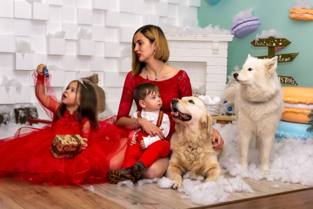 Family photo portrait. Mom and her two children and two white dogs in red clothes celebrate the Chistmas, new year. Happy young mother and her daughter, son and dogs in a cozy dark living room on Christmas eve. Celebrating Xmas at home. Winter evening with family, kids and dogs. A little girl in a red dress is holding a gift. Family looking at camera holding many gifts boxes celebrating happy New Year. Merry Christmas presents. A child in a red dress holds a gift box.Christmas conceptの写真素材