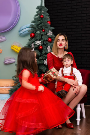 Family photo portrait. Mom and her two children in red clothes celebrate the Chistmas, new year. Happy young mother and her daughter and son in a cozy dark living room on Christmas eve. Celebrating Xmas at home. Winter evening with family and kids. A little girl in a red dress is holding a gift. Family looking at camera holding many gifts boxes celebrating happy New Year. Merry Christmas presents. A child in a red dress holds a gift box.Christmas concept. High quality photoの写真素材