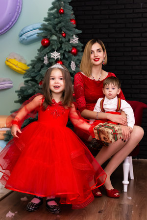 Family photo portrait. Mom and her two children in red clothes celebrate the Chistmas, new year. Happy young mother and her daughter and son in a cozy dark living room on Christmas eve. Celebrating Xmas at home. Winter evening with family and kids. A little girl in a red dress is holding a gift. Family looking at camera holding many gifts boxes celebrating happy New Year. Merry Christmas presents. A child in a red dress holds a gift box.Christmas concept. High quality photoの写真素材