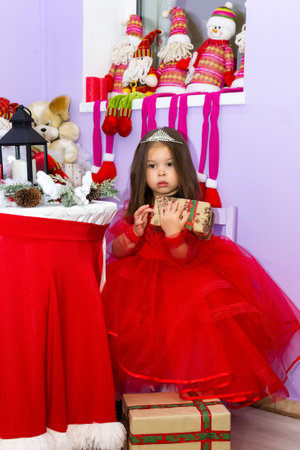 A little girl in a red dress is holding a gift. A little girl holds a gift on a chair. A child in a red dress holds a gift box. Girl looking at camera holding many gifts boxes celebrating happy New Year. Merry Christmas presents. Christmas concept. High quality photoの写真素材