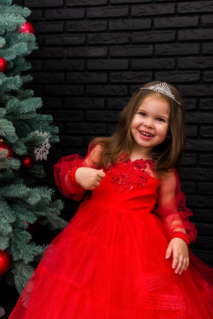 Little girl in red dress smiling by Christmas tree. Little beautiful girl in a red evening dress the Christmas tree. Girl in red in Christmas decorations. Christmas concept. New Year. Baby near a Christmas tree. High quality photoの写真素材