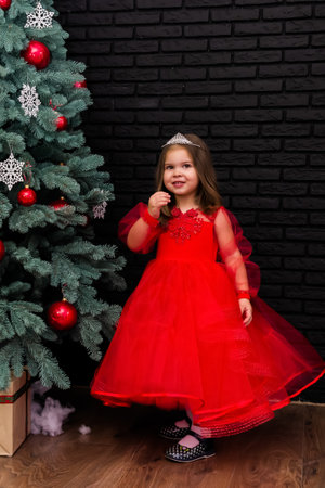 Little girl in red dress smiling by Christmas tree. Little beautiful girl in a red evening dress the Christmas tree. Girl in red in Christmas decorations. Christmas concept. New Year. Baby near a Christmas tree. High quality photoの写真素材