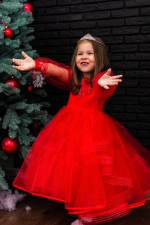 Little girl in red dress smiling by Christmas tree. Little beautiful girl in a red evening dress the Christmas tree. Girl in red in Christmas decorations. Christmas concept. New Year. Baby near a Christmas tree. High quality photoの写真素材