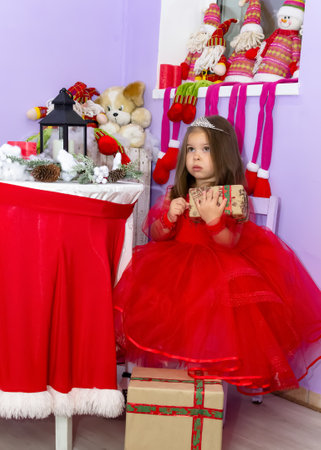 A little girl in a red dress is holding a gift. A little girl holds a gift on a chair. A child in a red dress holds a gift box. Girl looking at camera holding many gifts boxes celebrating happy New Year. Merry Christmas presents. Christmas concept. High quality photoの写真素材