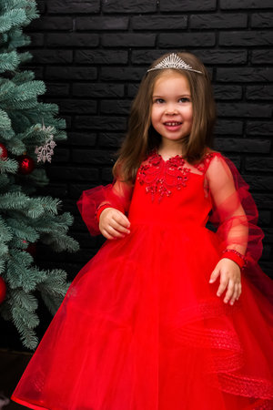 Little girl in red dress smiling by Christmas tree. Little beautiful girl in a red evening dress the Christmas tree. Girl in red in Christmas decorations. Christmas concept. New Year. Baby near a Christmas tree. High quality photoの写真素材