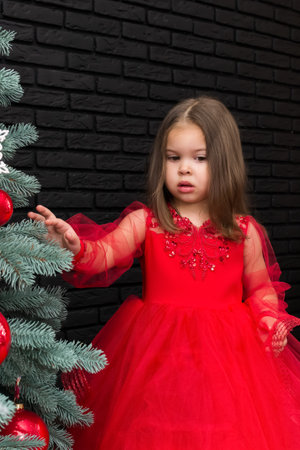 Little girl in red dress smiling by Christmas tree. Little beautiful girl in a red evening dress the Christmas tree. Girl in red in Christmas decorations. Christmas concept. New Year. Baby near a Christmas tree. High quality photoの写真素材