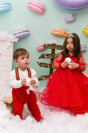 A little girl and a boy in red clothes have fun in the artificial snow by the fireplace in the studio. The girl is holding a box with gifts. Little kids in red christmas costume having fun beside a decorated christmas tree. Two kids playing with falling artificial snow flakes near a christmas tree.Kids playing with artificial snow flakes falling near a christmas tree. Christmas concept. High quality photoの写真素材