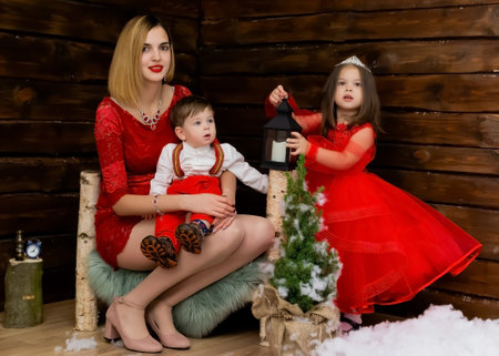 A little girl and a boy and their mother look at the Christmas candle. Wooden background. Preparing for the New Year and Christmas. A cheerful little girl, boy and woman looks at the Christmas lantern with a smile. Brother and sister and their mom in the red clothes at Christmas. Christmas concept. High quality photoの写真素材