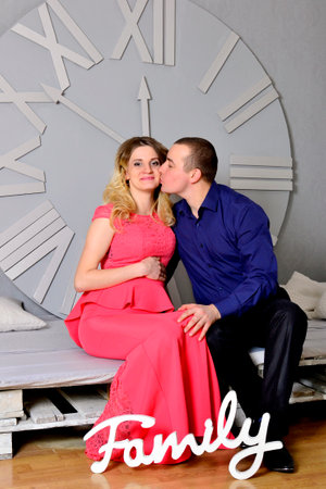 Attractive young couple is celebrating holiday at home. Standing on the background of beautiful Christmas tree husband hugs his pregnant wife. A man and his pregnant wife stand by a clock. New Years clock on the wall. High quality photoの写真素材