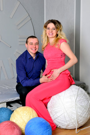 Attractive young couple is celebrating holiday at home. Standing on the background of beautiful Christmas tree husband hugs his pregnant wife. A man and his pregnant wife stand by a Christmas tree. Clock. New Years clock on the wall. High quality photoの写真素材