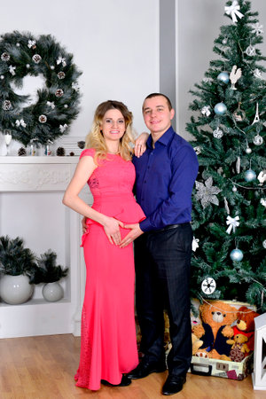 Attractive young couple is celebrating holiday at home. Standing on the background of beautiful Christmas tree husband hugs his pregnant wife. A man and his pregnant wife stand by a Christmas tree. Clock. New Years clock on the wall. High quality photoの写真素材
