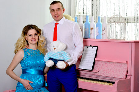 Attractive young couple is celebrating holiday at home. Standing on the background of beautiful a pink piano. Husband hugs his pregnant wife. A man and his pregnant wife stand by a piano. Standing and smiling near a pink piano. High quality photoの写真素材