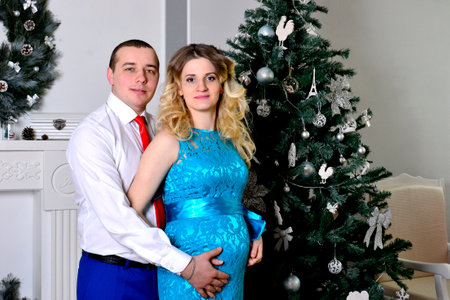 Attractive young couple is celebrating holiday at home. Standing on the background of beautiful Christmas tree husband hugs his pregnant wife. A man and his pregnant wife stand by a Christmas tree. High quality photoの写真素材