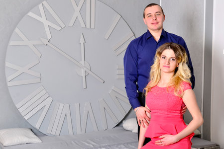 Attractive young couple is celebrating holiday at home. Standing on the background of beautiful Christmas tree husband hugs his pregnant wife. A man and his pregnant wife stand by a clock. New Years clock on the wall. High quality photoの写真素材