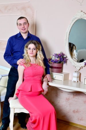 Attractive young couple is celebrating holiday at home. Standing and sitting on the background of a mirror. Husband hugs his pregnant wife. Family standing and sitting near a table with a mirror. High quality photoの写真素材