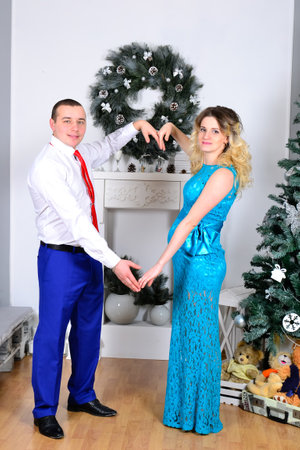 Attractive young couple is celebrating holiday at home. Standing on the background of beautiful Christmas tree husband hugs his pregnant wife. A man and his pregnant wife stand by a Christmas tree. High quality photoの写真素材