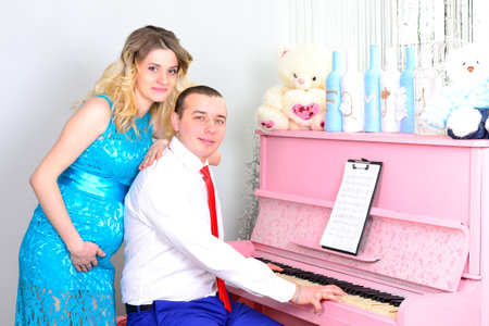 Attractive young couple is celebrating holiday at home. Standing on the background of beautiful a pink piano. Husband hugs his pregnant wife. A man and his pregnant wife stand by a piano. Standing and smiling near a pink piano. High quality photoの写真素材