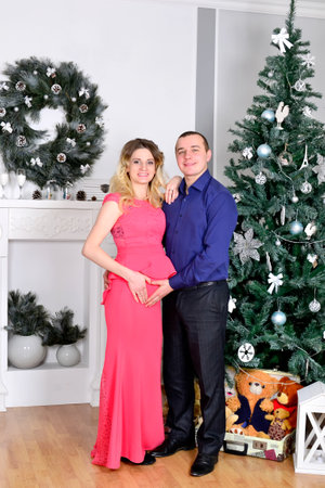Attractive young couple is celebrating holiday at home. Standing on the background of beautiful Christmas tree husband hugs his pregnant wife. A man and his pregnant wife stand by a Christmas tree. High quality photoの写真素材