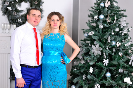 Attractive young couple is celebrating holiday at home. Standing on the background of beautiful Christmas tree husband hugs his pregnant wife. A man and his pregnant wife stand by a Christmas tree. High quality photoの写真素材