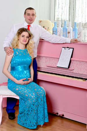 Attractive young couple is celebrating holiday at home. Standing on the background of beautiful a pink piano. Husband hugs his pregnant wife. A man and his pregnant wife stand by a piano. Standing and smiling near a pink piano. High quality photoの写真素材