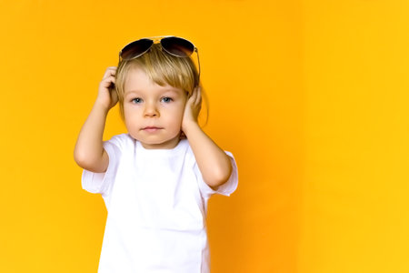 A 2-year-old child with blond hair wearing a white t-shirt with sunglasses on a yellow background. Emotions. The concept of childhood and family. Child front view on yellow isolated background with happy and excited expressionの写真素材