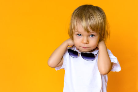 A 2-year-old child with blond hair wearing a white t-shirt with sunglasses on a yellow background. Emotions. The concept of childhood and family. Child front view on yellow isolated background with happy and excited expressionの写真素材