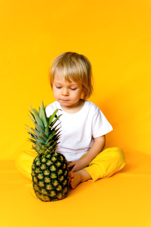 A 2-year-old child with blond hair in a white t-shirt and yellow pants with a ripe big pineapple on a yellow background. emotions The concept of childhood and family. Front view of child on yellow isolated background with happy and excited expression.の写真素材