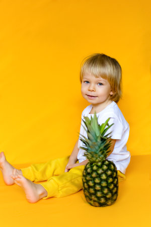 A 2-year-old child with blond hair in a white t-shirt and yellow pants with a ripe big pineapple on a yellow background. emotions The concept of childhood and family. Front view of child on yellow isolated background with happy and excited expression.の写真素材