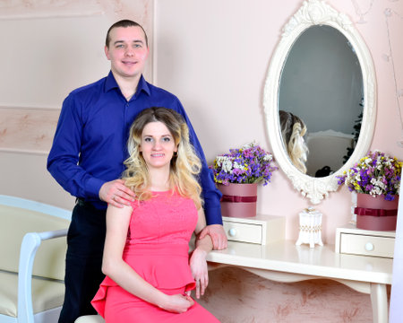 Attractive young couple is celebrating holiday at home. Standing and sitting on the background of a mirror. Husband hugs his pregnant wife. Family standing and sitting near a table with a mirror. High quality photoの写真素材