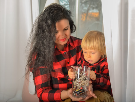 Mom and son dressed in identical festive Christmas clothes in a check sitting on the windowsill with a Christmas garland. Family look. Christmas miracle. Portrait of family mother with little son looking out window on Christmas Eve at home. Mom and kid sitting on windowsill with a Christmas decor on xmas eveningの写真素材
