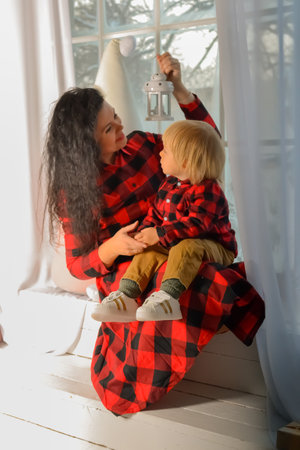 Mom and son dressed in identical festive Christmas clothes in a check sitting on the windowsill with a Christmas lantern. Family look. Christmas miracle. Portrait of family mother with little son looking out window on Christmas Eve at home. Mom and kid sitting on windowsill with glowing lantern on xmas eveningの写真素材