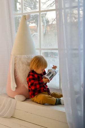 The kid sits on the windowsill with a Christmas gnome and holds a Christmas lantern and a New Years gift, box in his hands. Family Christmas Conceptの写真素材