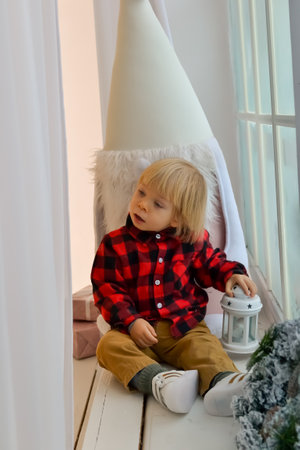 The kid sits on the windowsill with a Christmas gnome and holds a Christmas lantern and a New Years gift, box in his hands. Family Christmas Conceptの写真素材