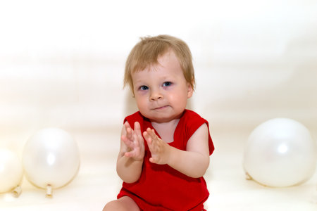 One-year-old toddler with blonde hair in red bodysuit sitting and smiling on white background with inflatable balloons. Wonderful kid sat down to rest. Baby posing isolated over light backgroundの写真素材