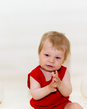 One-year-old toddler with blonde hair in red bodysuit sitting and smiling on white background with inflatable balloons. Wonderful kid sat down to rest. Baby posing isolated over light backgroundの写真素材