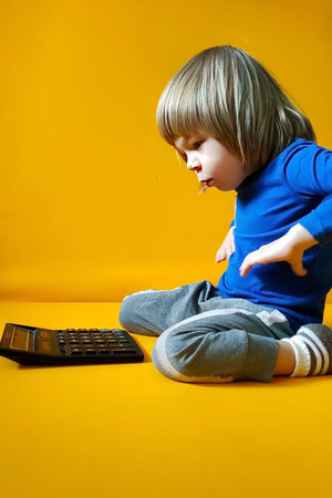 A small child counts on a calculator on a yellow background. Kid fingers press buttons and calculate studying math at home. Child makes arithmetic calculations with a calculatorの写真素材