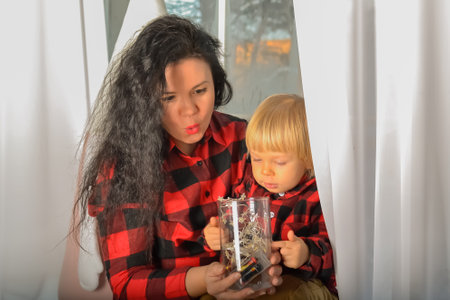 Mom and son dressed in identical festive Christmas clothes in a check sitting on the windowsill with a Christmas garland. Family look. Christmas miracle. Portrait of family mother with little son looking out window on Christmas Eve at home. Mom and kid sitting on windowsill with a Christmas decor on xmas eveningの写真素材