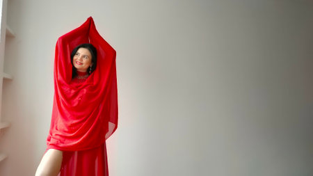 A dancer in a red suit is dancing with a red shawl an oriental dance on a white background. Free space for text. Banner. A professional dancer dances oriental belly dance. Sexy woman in red dress dances with a red shawl a seductive dance in a white studio. Isolation. Professional dance trainerの写真素材