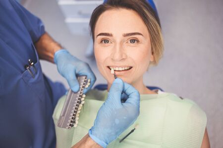 Dentist checks the level of patient's teeth whitening with a dentist's color. Dental equipment in dentistry office. Stomatology concept. Doctor's hands in medical gloves and smiling happy woman.の写真素材