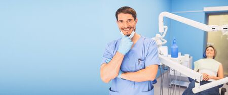 Young smiling handsome male doctor in blue medical uniform, disposable medical facial mask with equipment in dentistry office and happy patient in the dental chair. Stomatology concept. Copy spaceの写真素材