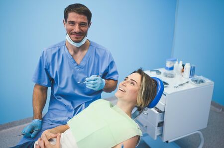 Dentist examining a patient's teeth using dental equipment in dentistry office. Stomatology and health care concept. Young handsome male doctor in disposable medical facial mask, smiling happy woman.の写真素材