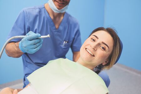 Dentist examining a patients teeth using dental equipment in dentistry office. Stomatology and health concept. Young handsome male doctor in disposable medical facial mask, smiling happy woman.の写真素材